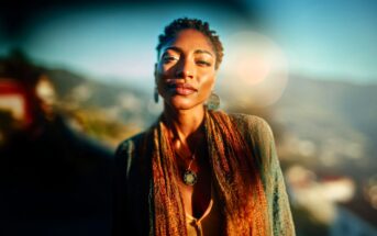 A woman with short braided hair, wearing a patterned shawl and pendant necklace, stands outdoors in sunlight with a blurred landscape and lens flare in the background.