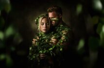 A man and woman are wrapped in green vines and leaves, standing close together in an embrace. The scene is dimly lit, with a dark background and a dreamy, nature-inspired atmosphere.