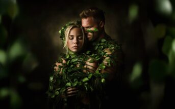 A man and woman are wrapped in green vines and leaves, standing close together in an embrace. The scene is dimly lit, with a dark background and a dreamy, nature-inspired atmosphere.