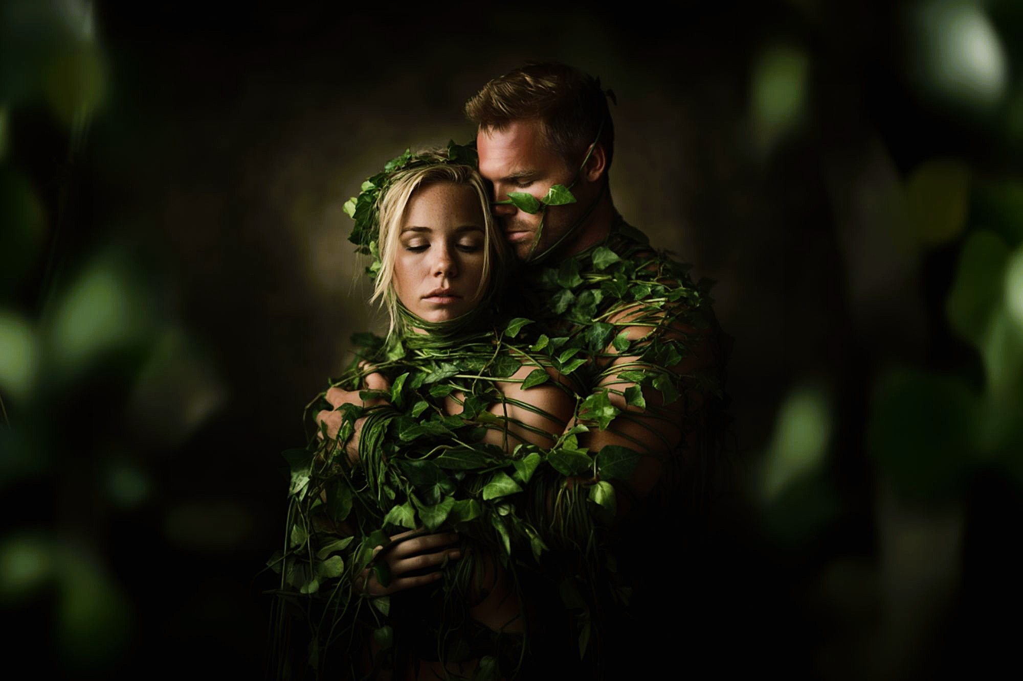 A man and woman are wrapped in green vines and leaves, standing close together in an embrace. The scene is dimly lit, with a dark background and a dreamy, nature-inspired atmosphere.