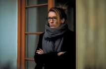 A woman with glasses and a large gray scarf stands with her arms crossed, looking serious. She is in front of a glass door with a wooden frame, and the background is softly blurred.