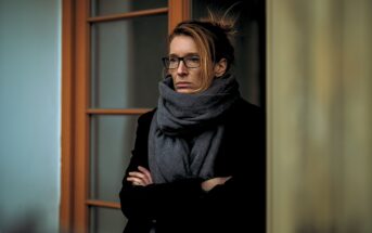 A woman with glasses and a large gray scarf stands with her arms crossed, looking serious. She is in front of a glass door with a wooden frame, and the background is softly blurred.