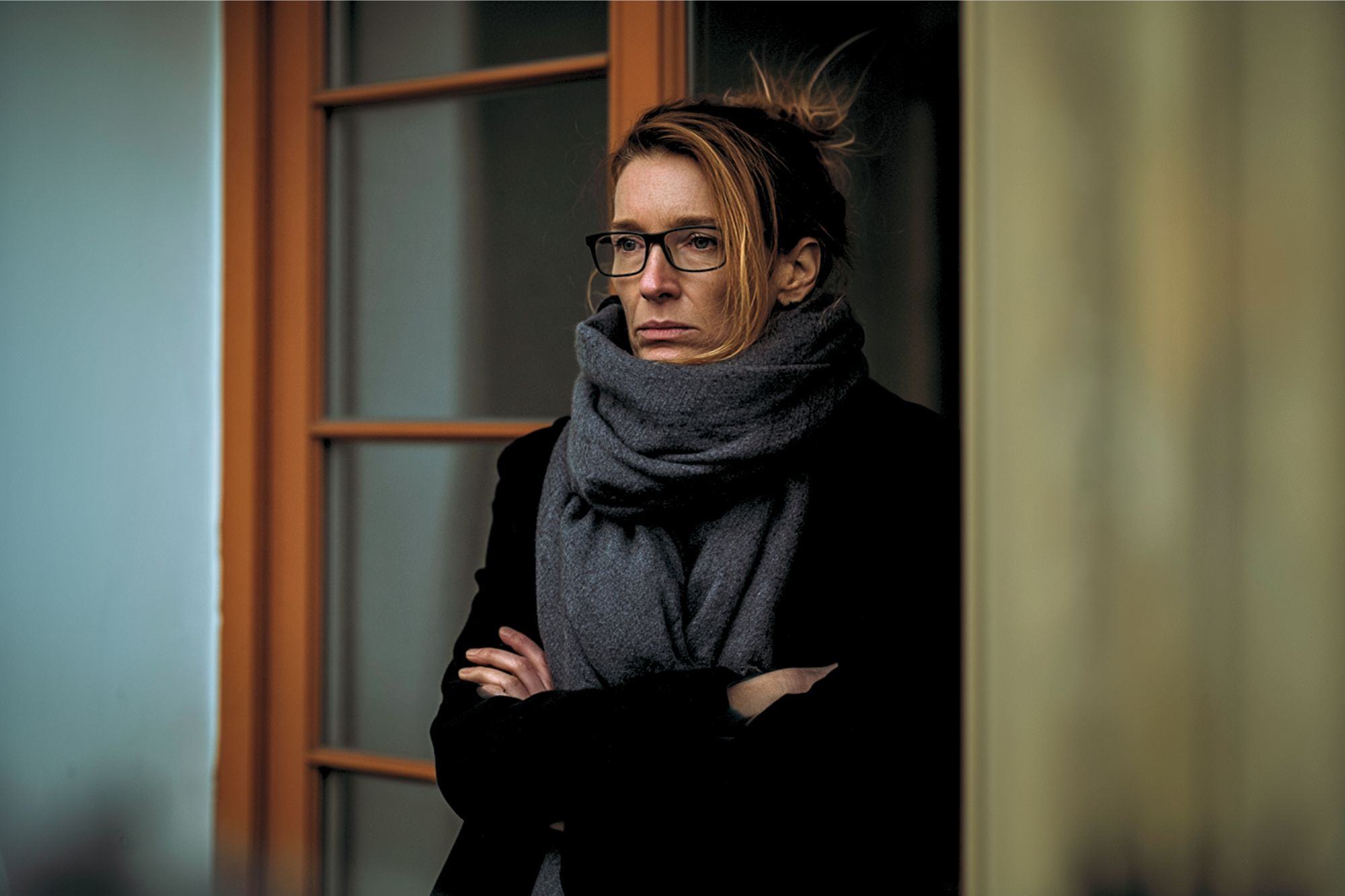 A woman with glasses and a large gray scarf stands with her arms crossed, looking serious. She is in front of a glass door with a wooden frame, and the background is softly blurred.