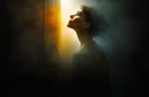 A woman stands in a dark, smoky room, gazing upward toward a bright, warm light streaming through a slightly open door, illuminating her face and silhouette.