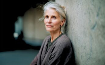 An older woman with gray hair and a calm expression leans against a textured wall, wearing a gray cardigan. The background is softly blurred, highlighting her gentle, contemplative demeanor.