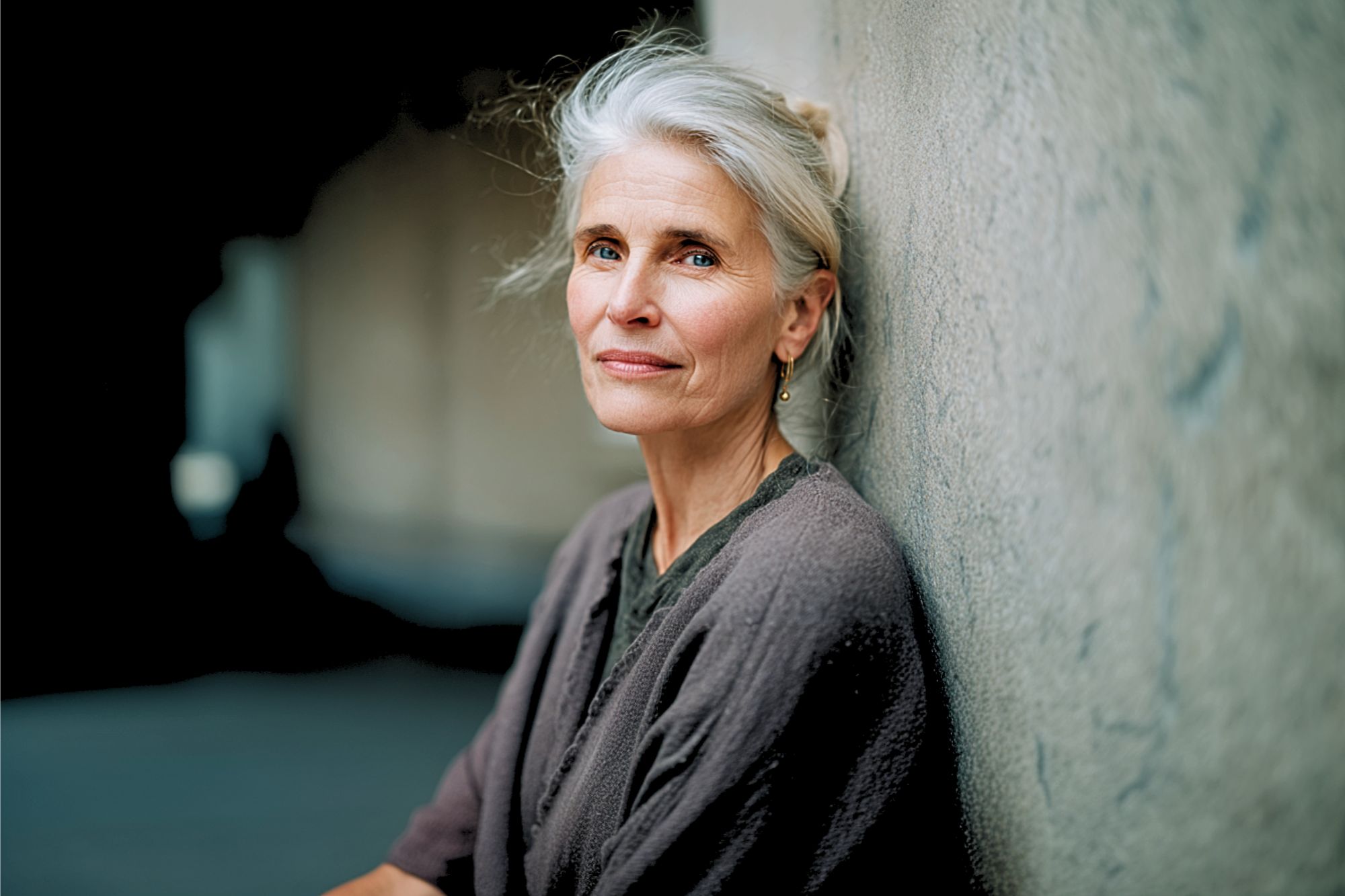 An older woman with gray hair and a calm expression leans against a textured wall, wearing a gray cardigan. The background is softly blurred, highlighting her gentle, contemplative demeanor.