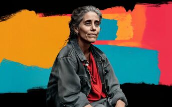 A woman with gray-streaked hair wearing a dark denim jacket and red shirt sits against a background of bold, abstract colors in orange, blue, and red brushstrokes.