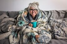 A smiling person with gray hair and glasses sits on a couch, wrapped in a floral blanket and holding a turquoise mug, surrounded by cozy pillows and throws.