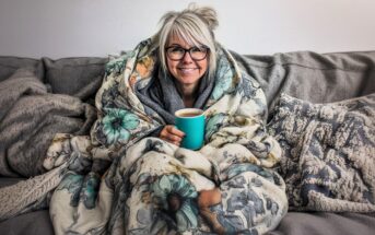 A smiling person with gray hair and glasses sits on a couch, wrapped in a floral blanket and holding a turquoise mug, surrounded by cozy pillows and throws.