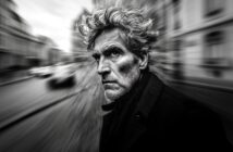 Black and white portrait of a man with wild hair and a serious expression, set against a blurred urban background.