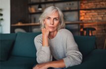 A woman with gray hair and a light sweater sits on a teal couch, resting her chin on her hand and looking thoughtfully at the camera. The room has warm lighting and shelves in the background.