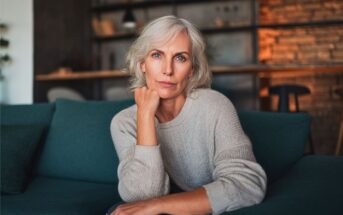 A woman with gray hair and a light sweater sits on a teal couch, resting her chin on her hand and looking thoughtfully at the camera. The room has warm lighting and shelves in the background.
