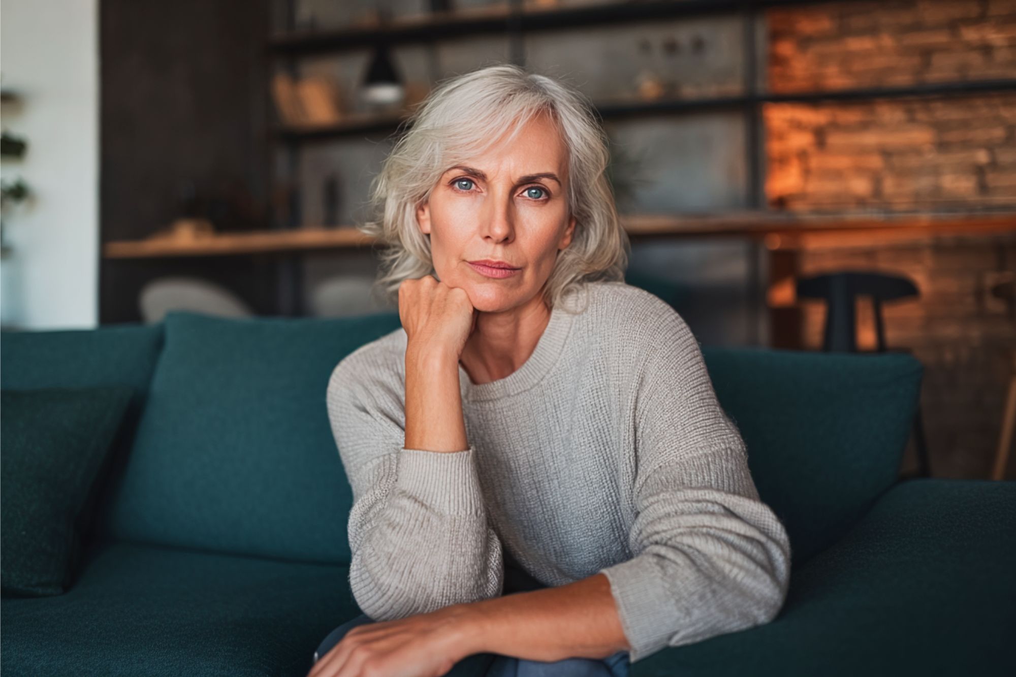 A woman with gray hair and a light sweater sits on a teal couch, resting her chin on her hand and looking thoughtfully at the camera. The room has warm lighting and shelves in the background.