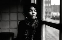A woman with curly hair and hoop earrings sits indoors by a window, gazing thoughtfully outside; the scene is in black and white, with soft natural light illuminating her face.