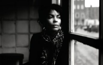 A woman with curly hair and hoop earrings sits indoors by a window, gazing thoughtfully outside; the scene is in black and white, with soft natural light illuminating her face.