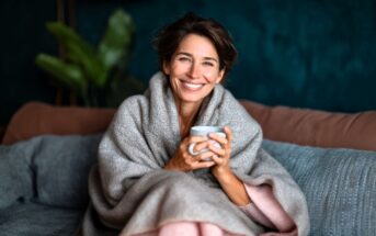 A woman with short brown hair, wrapped in a cozy gray blanket, sits on a sofa holding a mug and smiling warmly. The background features a dark teal wall and green indoor plant.