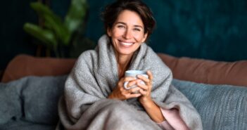 A woman with short brown hair, wrapped in a cozy gray blanket, sits on a sofa holding a mug and smiling warmly. The background features a dark teal wall and green indoor plant.