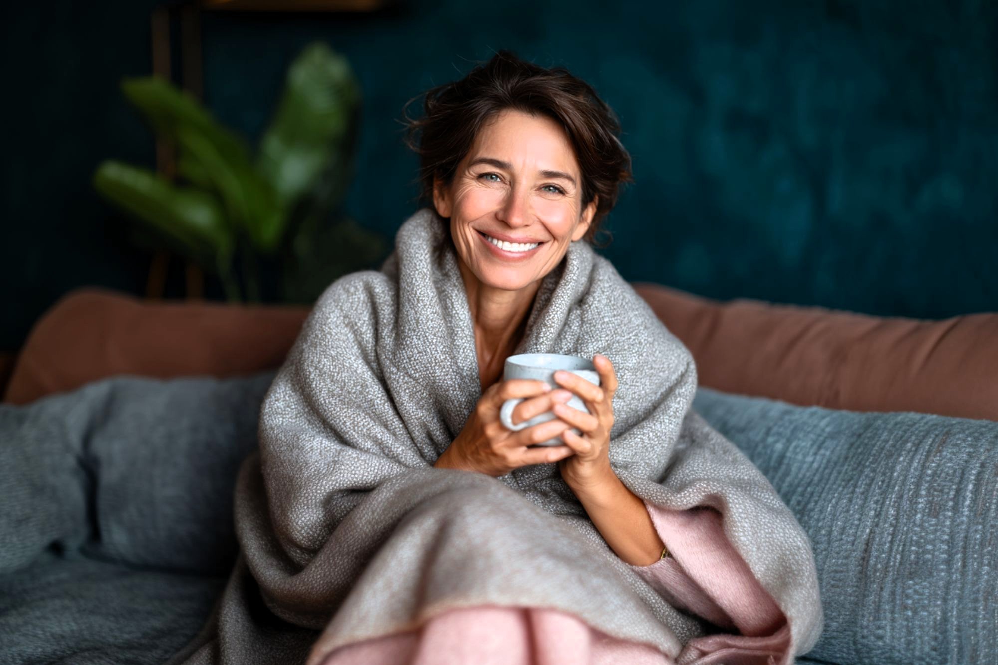 A woman with short brown hair, wrapped in a cozy gray blanket, sits on a sofa holding a mug and smiling warmly. The background features a dark teal wall and green indoor plant.