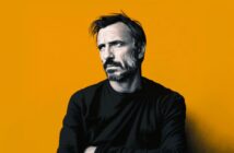 A man with short dark hair, a beard, and a serious expression is wearing a black shirt and standing against a solid orange background.