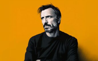 A man with short dark hair, a beard, and a serious expression is wearing a black shirt and standing against a solid orange background.