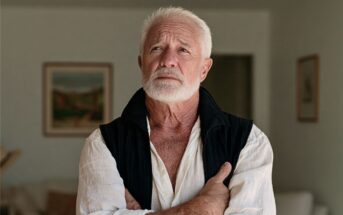 An elderly man with white hair and a beard stands indoors with arms crossed, wearing a white shirt and dark vest, looking thoughtfully upward. Paintings hang on the wall behind him.