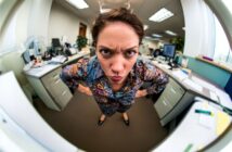A woman stands in an office with her hands on her hips, leaning forward and frowning at the camera. The image is taken with a fisheye lens, causing a distorted, wide-angle effect.