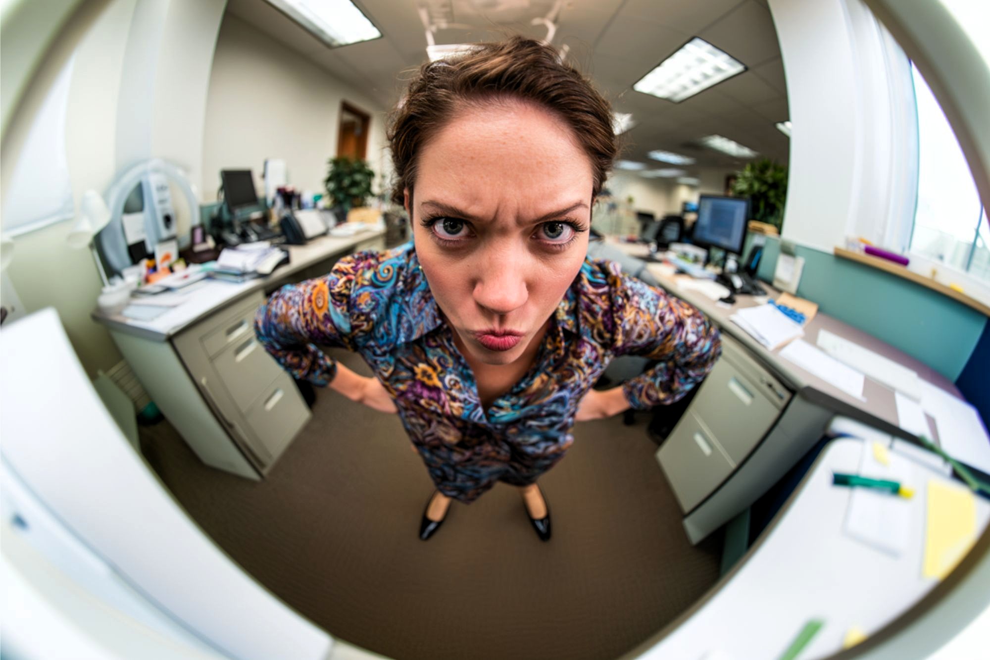 A woman stands in an office with her hands on her hips, leaning forward and frowning at the camera. The image is taken with a fisheye lens, causing a distorted, wide-angle effect.