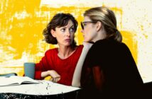 Two women sit at a table with an open notebook and a cup, engaged in a serious conversation. One woman in red looks concerned, while the other listens intently. The background is yellow with artistic splashes.