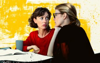 Two women sit at a table with an open notebook and a cup, engaged in a serious conversation. One woman in red looks concerned, while the other listens intently. The background is yellow with artistic splashes.