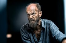 An older man with a wrinkled forehead and bushy beard glares intensely, showing clenched teeth and anger. He wears a rumpled blue shirt and is set against a dark, blurred background.