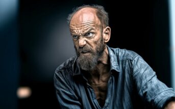 An older man with a wrinkled forehead and bushy beard glares intensely, showing clenched teeth and anger. He wears a rumpled blue shirt and is set against a dark, blurred background.