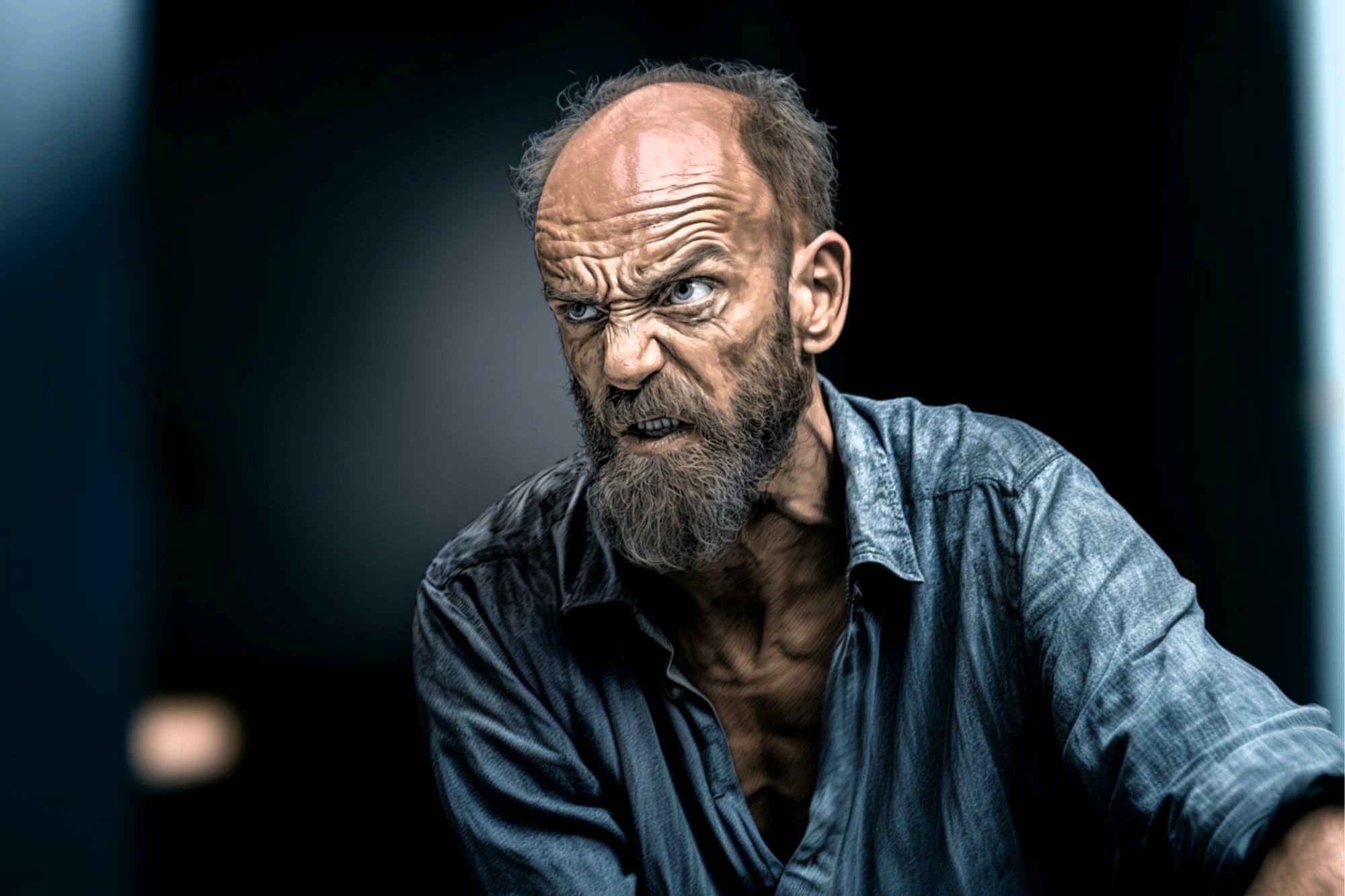 An older man with a wrinkled forehead and bushy beard glares intensely, showing clenched teeth and anger. He wears a rumpled blue shirt and is set against a dark, blurred background.