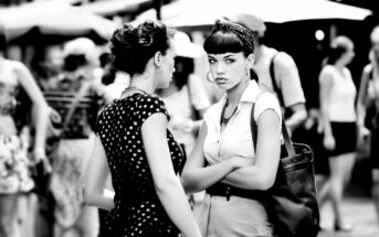 Two women stand close together in a busy outdoor setting. One faces the camera with arms crossed and a serious expression, while the other, seen from the side, appears to be talking. The background is blurred with people and umbrellas.