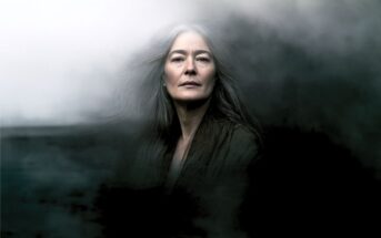 An older woman with long gray hair stands surrounded by swirling dark mist, looking directly at the camera with a calm, serious expression.