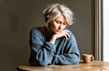 An older woman with short gray hair wearing a blue sweater sits at a wooden table, looking down thoughtfully with her hand near her face. A beige mug is on the table beside her, and soft light comes from the right.