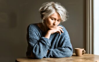 An older woman with short gray hair wearing a blue sweater sits at a wooden table, looking down thoughtfully with her hand near her face. A beige mug is on the table beside her, and soft light comes from the right.