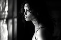 A woman with wavy hair gazes pensively out of a window, with dramatic light and shadow highlighting her face in a black and white setting.