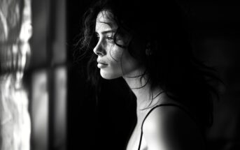 A woman with wavy hair gazes pensively out of a window, with dramatic light and shadow highlighting her face in a black and white setting.