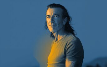 A middle-aged man with long hair and a beard stands outdoors, looking at the camera. The image has a blue and yellow tonal effect, with soft lighting highlighting his face and chest.