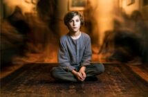 A young boy with brown hair sits cross-legged on a patterned rug, wearing loose gray clothes. The background is blurred, with shadowy, indistinct figures creating a sense of motion or ghostly presence around him.