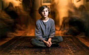 A young boy with brown hair sits cross-legged on a patterned rug, wearing loose gray clothes. The background is blurred, with shadowy, indistinct figures creating a sense of motion or ghostly presence around him.
