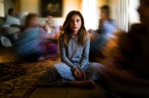 A young girl sits cross-legged on a wooden floor in a softly lit room, looking directly at the camera. The background and other children around her are blurred, creating a sense of motion and focus on her.