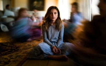 A young girl sits cross-legged on a wooden floor in a softly lit room, looking directly at the camera. The background and other children around her are blurred, creating a sense of motion and focus on her.