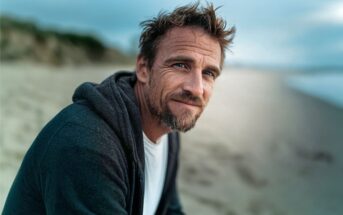 A man with tousled hair and a beard sits on a sandy beach, wearing a dark hoodie over a white shirt, looking at the camera with a soft smile. The background is blurred, showing sand and distant greenery.