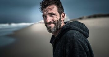 A middle-aged man with a beard and tousled hair, wearing a dark hoodie, stands on a sandy beach under a cloudy sky, smiling slightly at the camera. The ocean and dunes are blurred in the background.