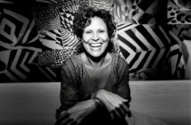 A woman with curly hair sits smiling broadly against a vibrant, geometric black-and-white background, her hands resting on her knees, exuding warmth and joy.