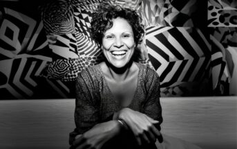 A woman with curly hair sits smiling broadly against a vibrant, geometric black-and-white background, her hands resting on her knees, exuding warmth and joy.