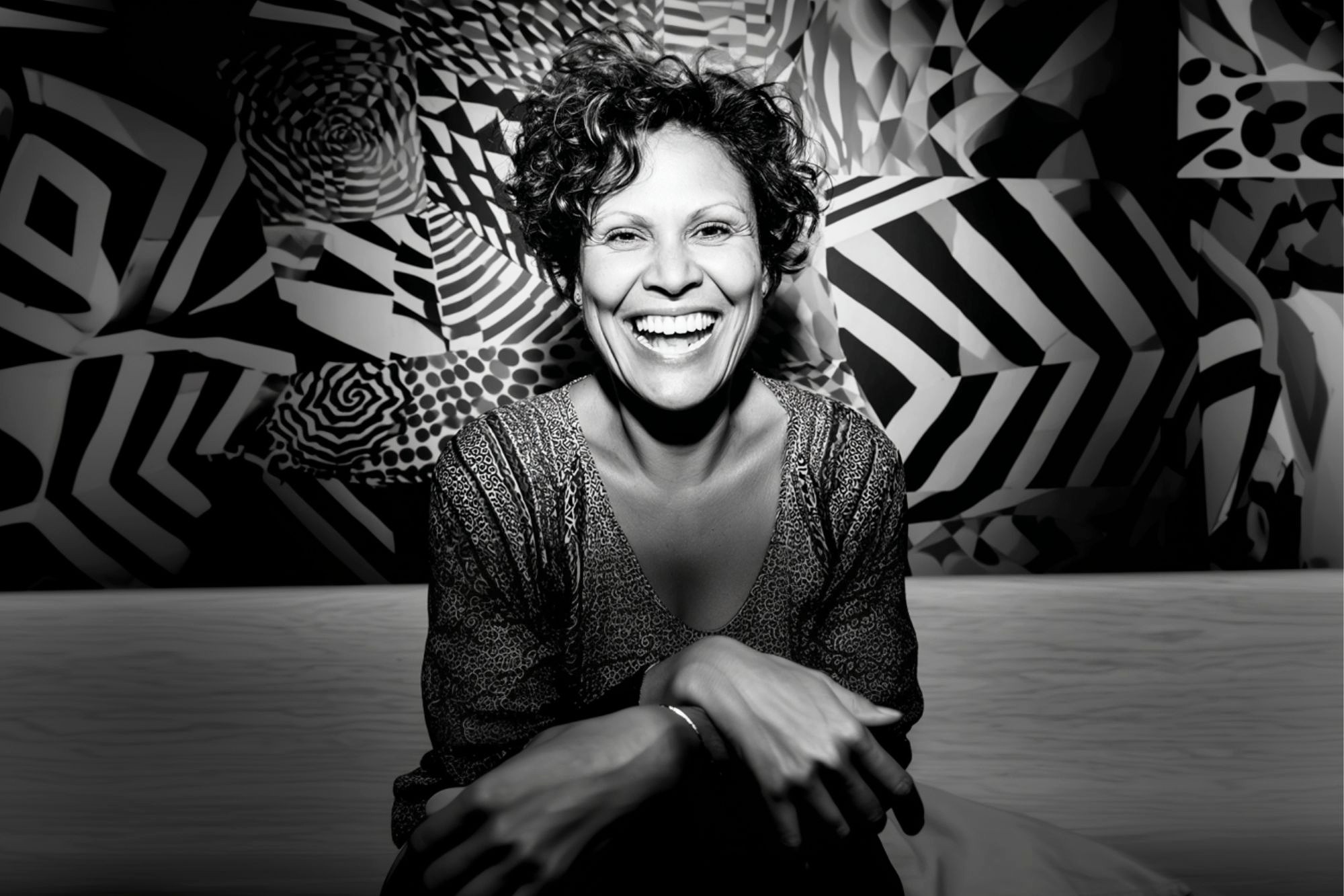 A woman with curly hair sits smiling broadly against a vibrant, geometric black-and-white background, her hands resting on her knees, exuding warmth and joy.