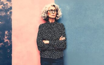 An older woman with curly gray hair, glasses, and a black polka dot shirt stands with arms crossed against a split pink and blue textured background.
