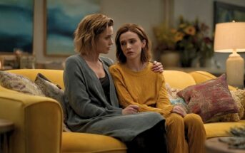 Two women sit close together on a yellow couch, one comforting the other with an arm around her shoulders. Both look serious and concerned. The room is warmly lit with art and flowers visible in the background.